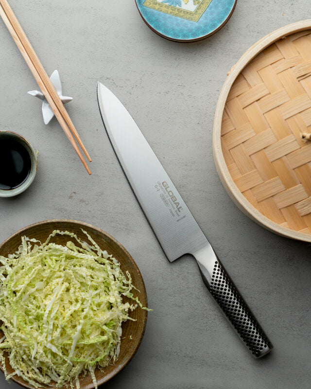 Global Japanese chefs knife, G-2 Cook's knife next to sliced cabbage, bamboo steamer and soy sauce. Professional stainless steel kitchen knife for chefs, vegetable prep and Asian cuisine cooking. Made in Japan.
