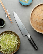 Global G-2 Japanese Chef's knife stainless steel  kitchen knife on a grey surface with shredded cabbage, chopsticks, and a bamboo steamer.