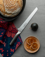 Global G-9 Bread knife with a serrated edge on a grey surface next to a loaf of bread, dried oranges, and a dark fruit. Bread knife for slicing sourdough read, home made bread and crusty bread.