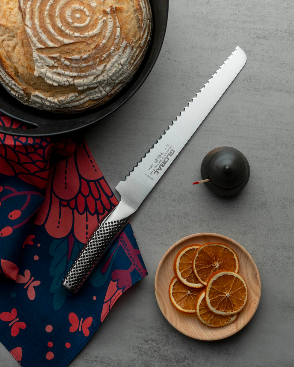Global G-9 Bread knife with a serrated edge on a grey surface next to a loaf of bread, dried oranges, and a dark fruit. Bread knife for slicing sourdough read, home made bread and crusty bread.