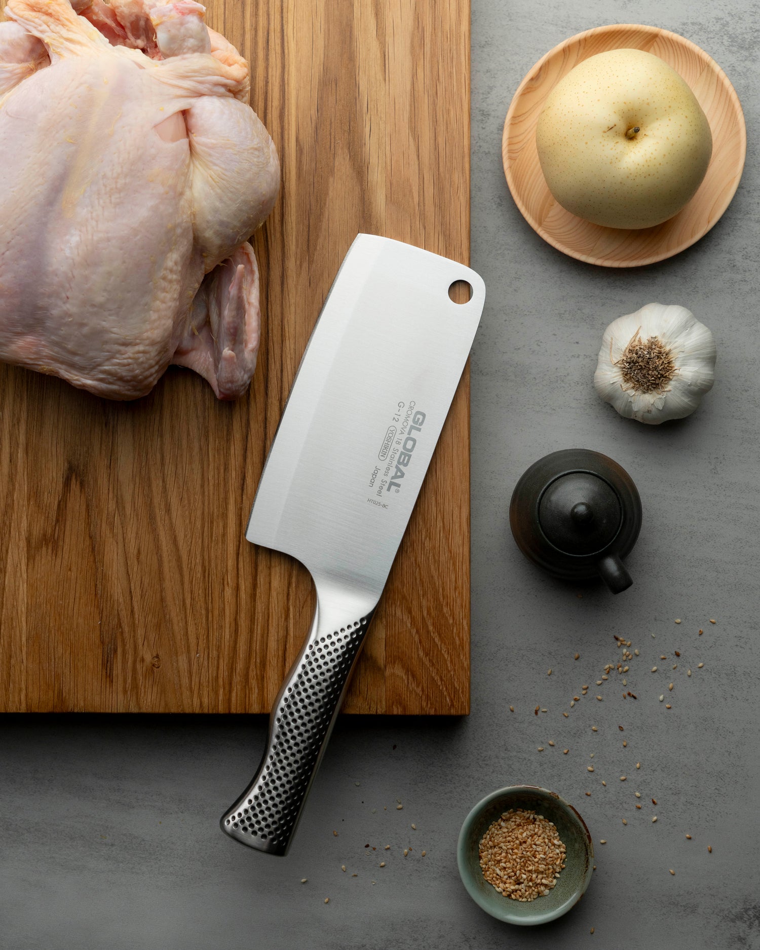 Global G-12 Meat cleaver on a wooden cutting board with a raw chicken, garlic, and spices on a gray surface.