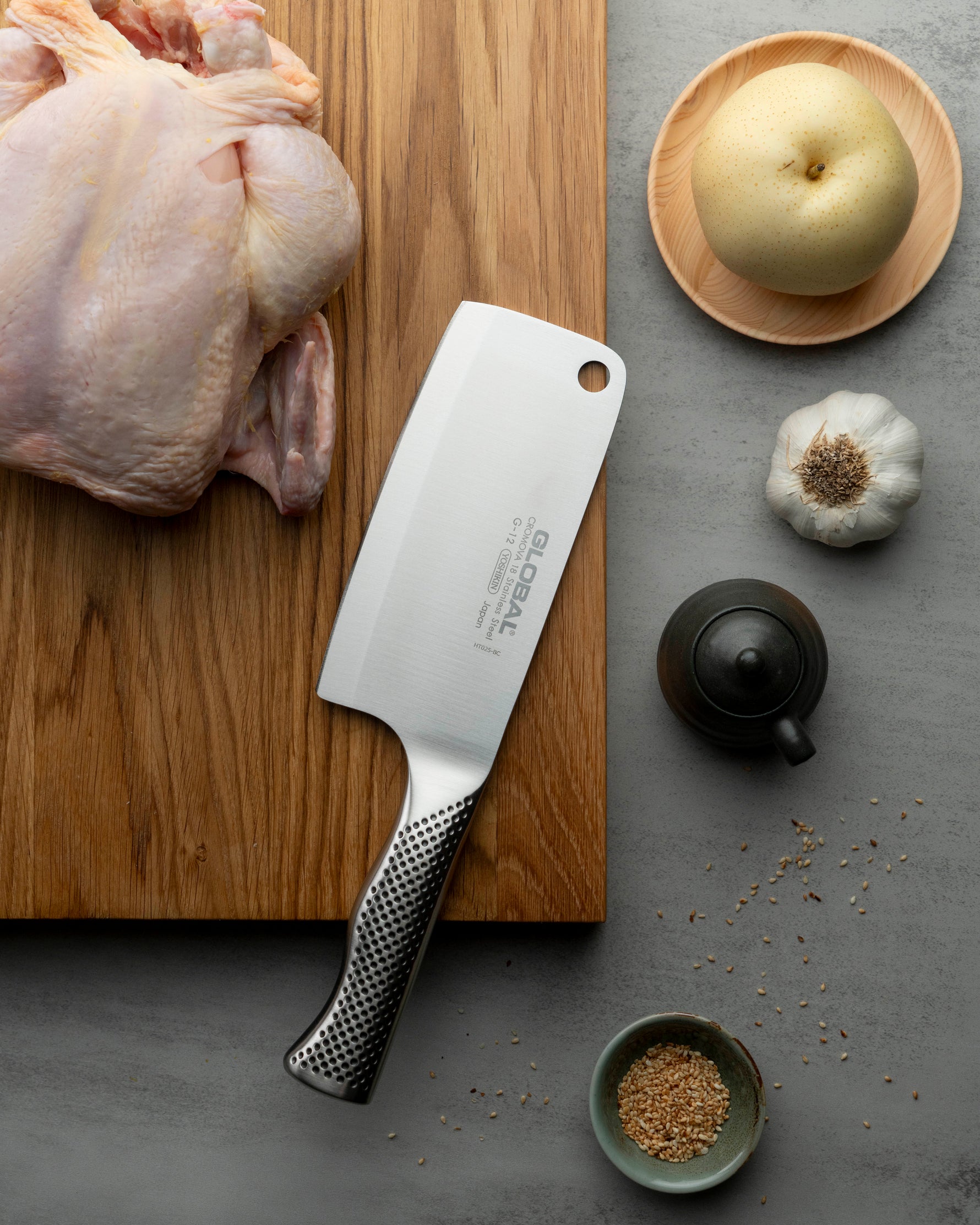Global G-12 Meat cleaver on a wooden cutting board with a raw chicken, garlic, and spices on a gray surface.