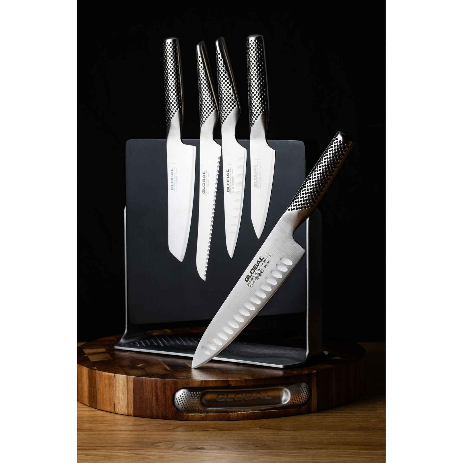 Set of kitchen knives on a wooden magnetic knife holder with a wooden base against a black background. 