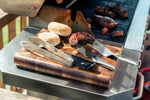 Global grilling tools and ingredients on a cutting board with a grill in the background. Stainless steel barbecue tools.