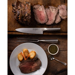 Sliced beef on a wooden cutting board with a  Global carving knife and fork set with plate of beef and vegetables, and a bowl of sauce.