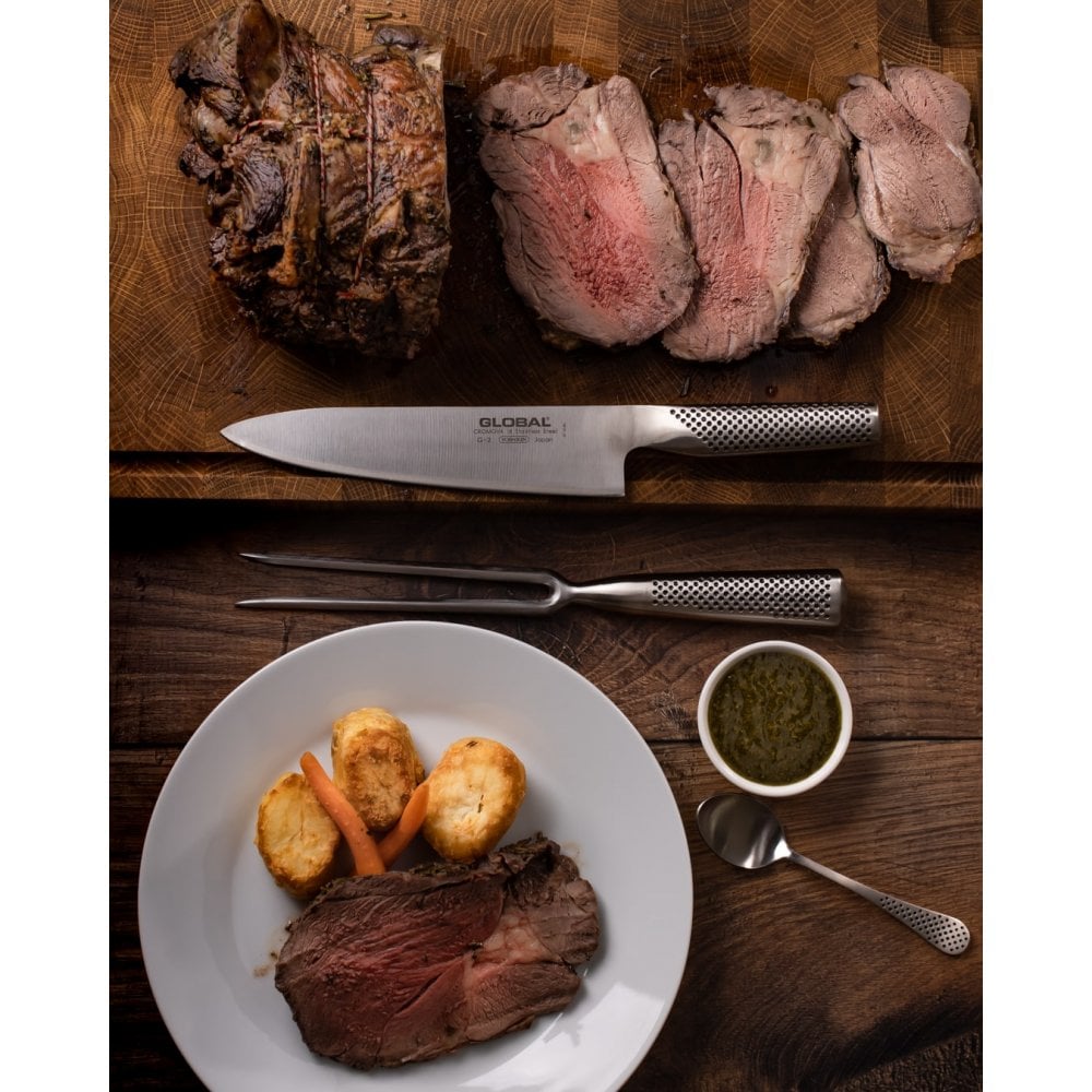 Sliced beef on a wooden cutting board with a  Global carving knife and fork set with plate of beef and vegetables, and a bowl of sauce.
