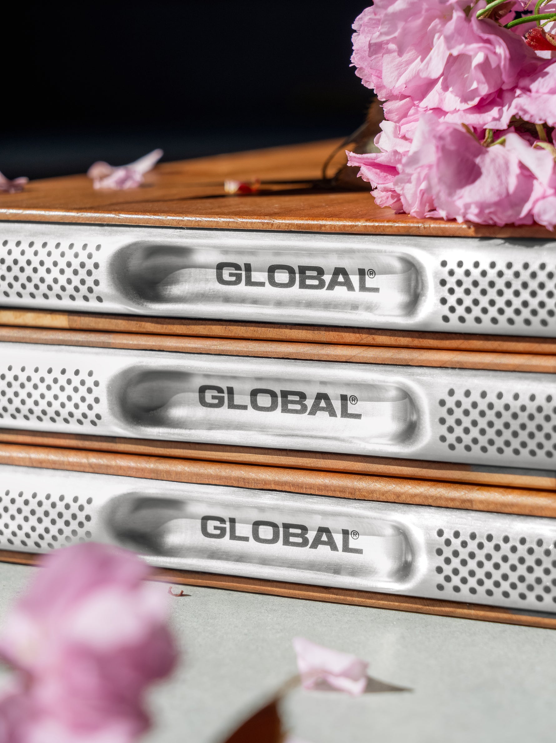 Close up of three GLOBAL premium wooden chopping boards made from cherry wood. Durable cutting boards with inbuilt metal handles with GLOBAL indentations for meal preparation on a surface with cherry blossom flowers.