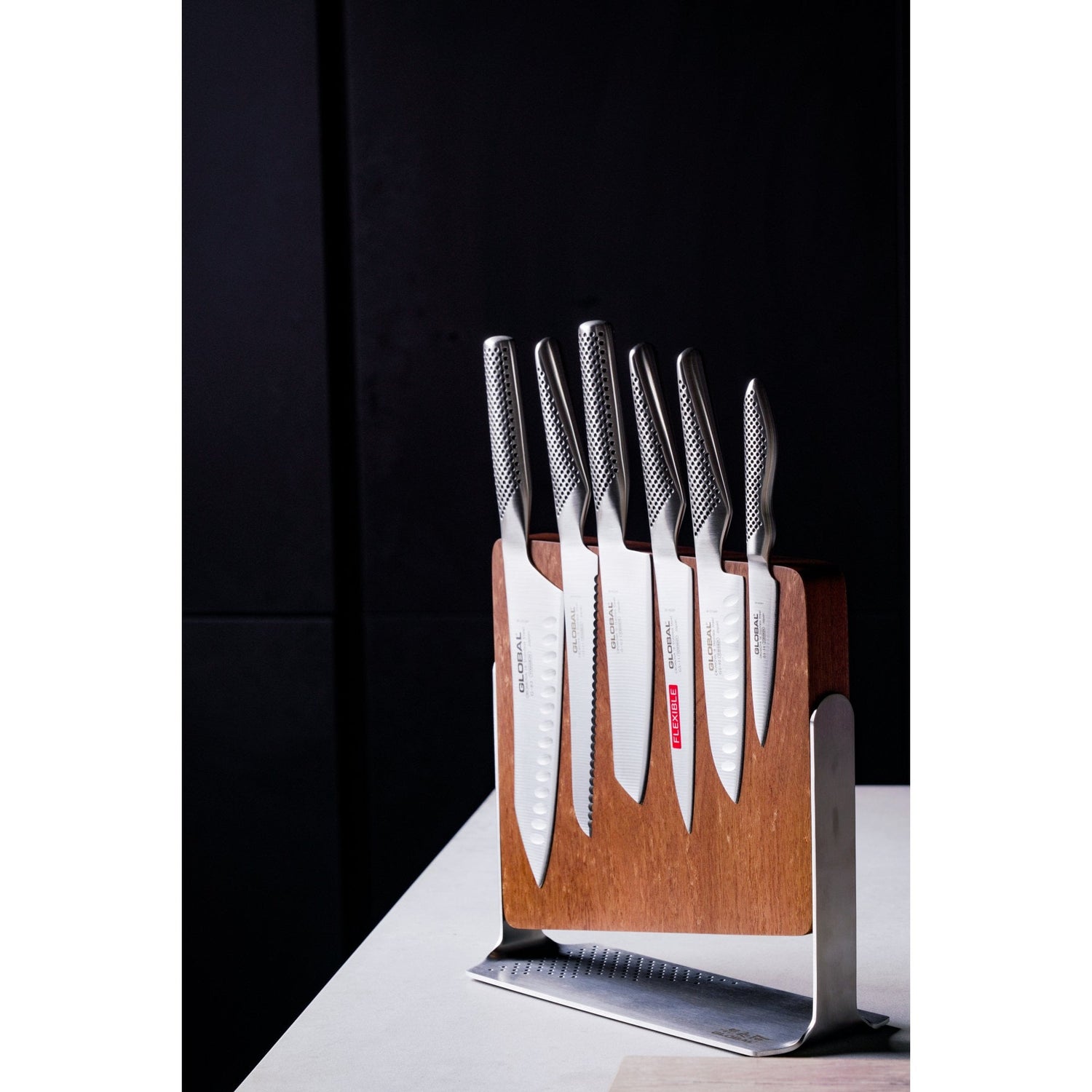 Set of Global knives on a wooden block with a black background. Magnetic knife block with cook's knife, vegetable chopping knife, utility knife, santoku knife, bread knife and paring knife.