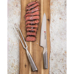 Sliced steak on a wooden cutting board with a Global carving knife and fork, on a textured surface.