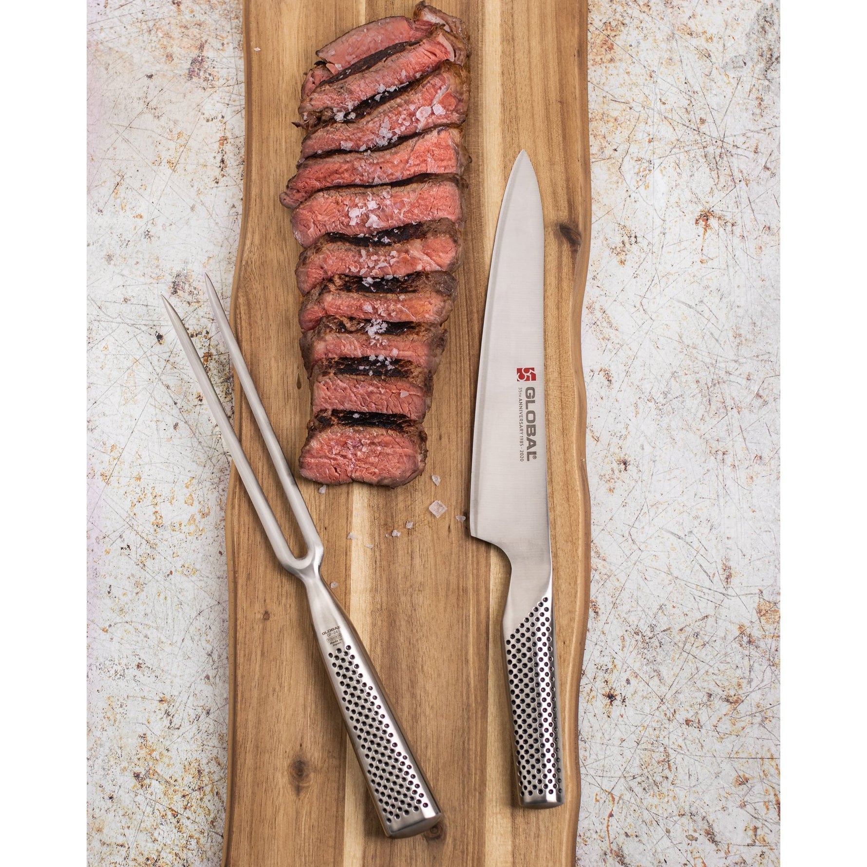 Sliced steak on a wooden cutting board with a Global carving knife and fork, on a textured surface.