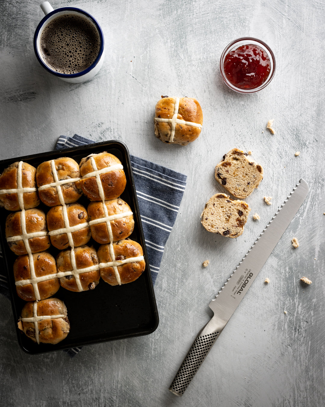 Hot cross buns on a tray with the global G-9 serrated kitchen knife, a bread knife, cup of coffee, and jar of jam on a grey surface.