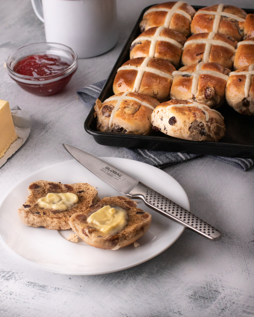 Hot cross buns on a tray with a plate of buns split open with butter, a sharp utility knife Global, and a jar of jam on a light gray surface.