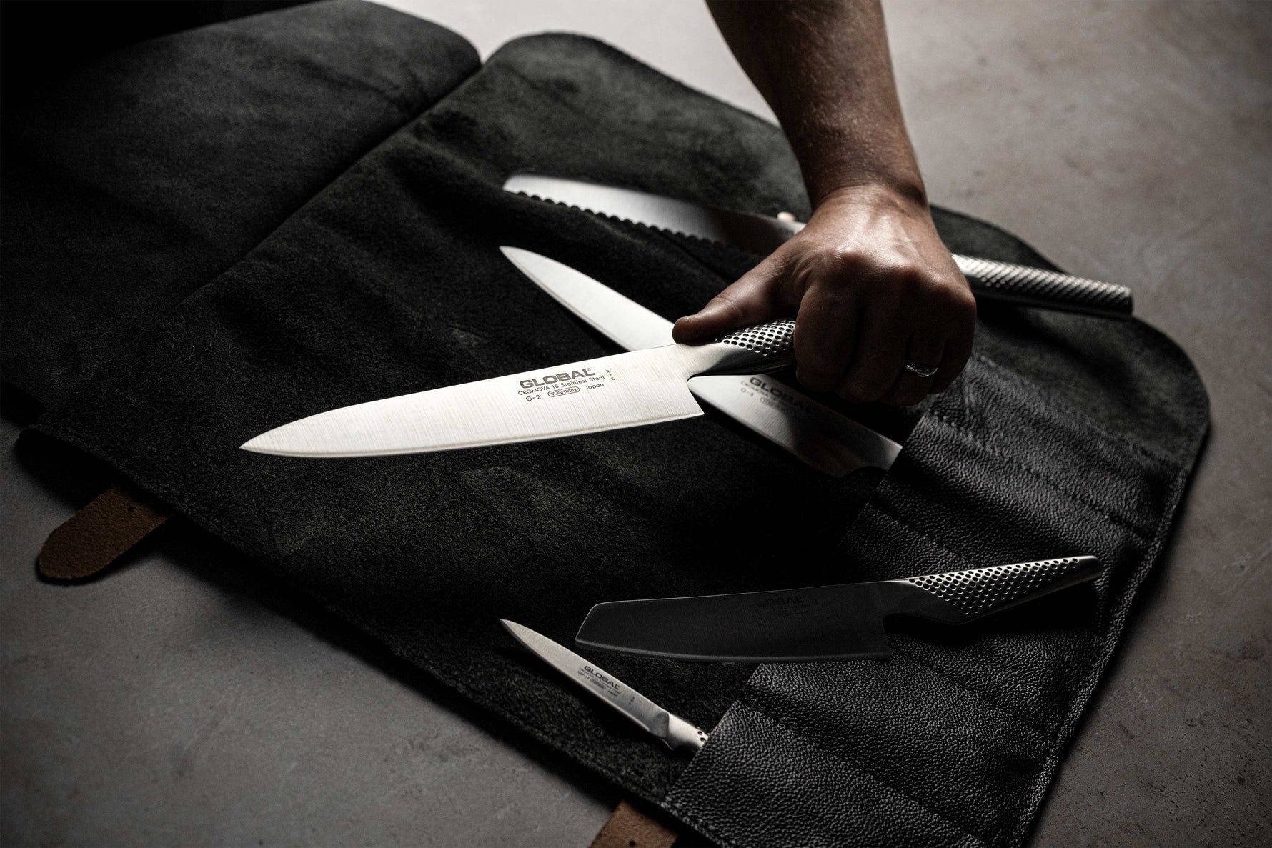 Chef holding Global G-2 chef's knife next to black leather knife roll case with 5 slots