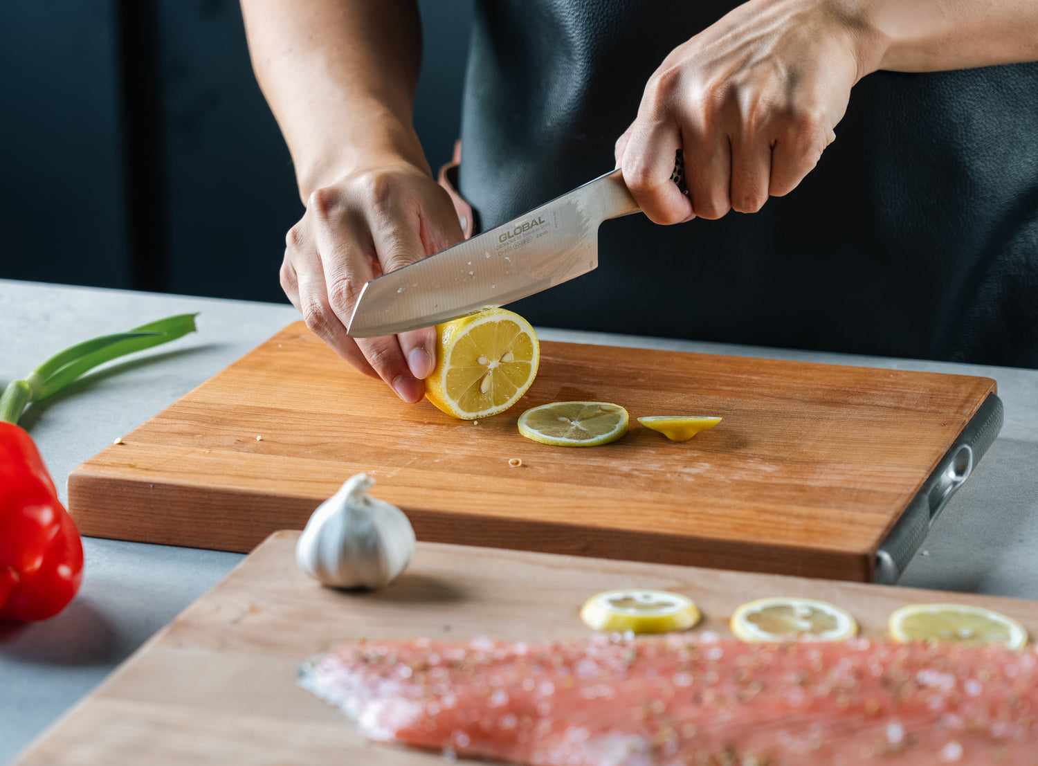 Professional chef cutting lemon, fish, garlic, spring onions and red pepper on Global wooden cutting board.