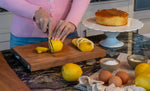Home baker cutting lemons on a Global cherry wood cutting board with a cake and eggs in the background. Home cook is using the global GSF-22 utility knife for slicing fruit.