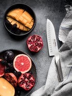 Fruits including pomegranate, grapefruit, and mango in bowls on a dark surface with a Global santoku knife, a Japanese chef's knife for cutting fruit, vegetable, meat and fish.