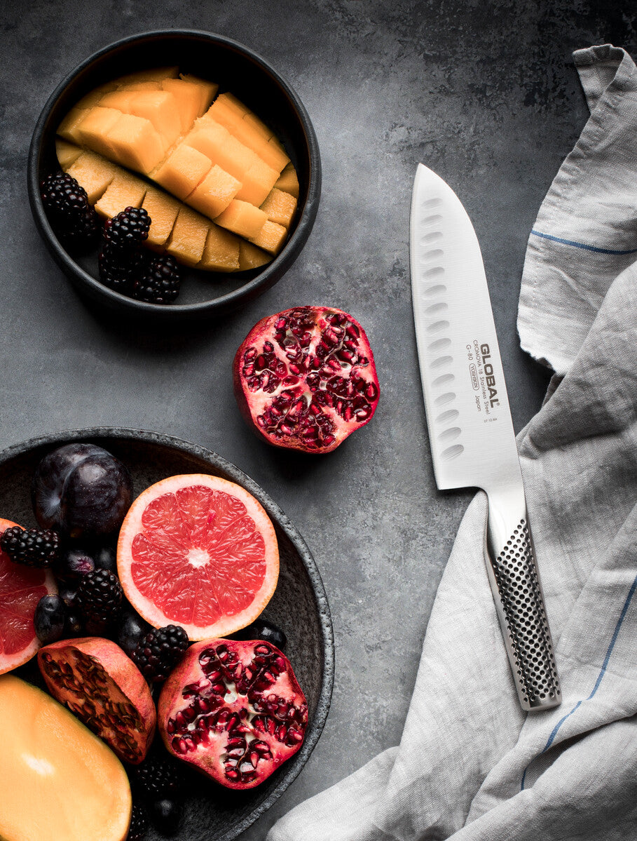 Fruits including pomegranate, grapefruit, and mango in bowls on a dark surface with a Global santoku knife, a Japanese chef's knife for cutting fruit, vegetable, meat and fish.