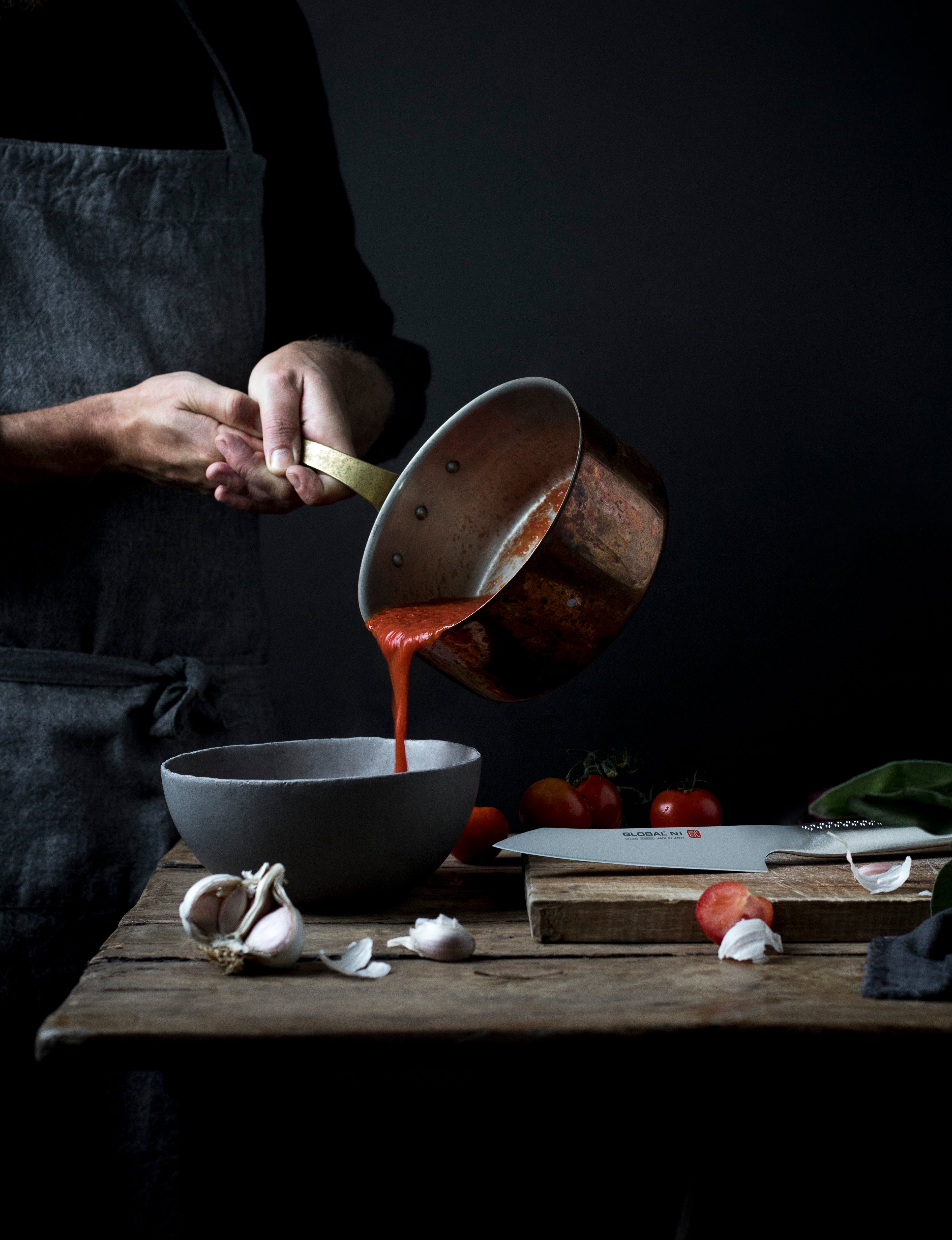 Person pouring tomato sauce from a pot into a bowl on a wooden table with ingredients and Global knife.