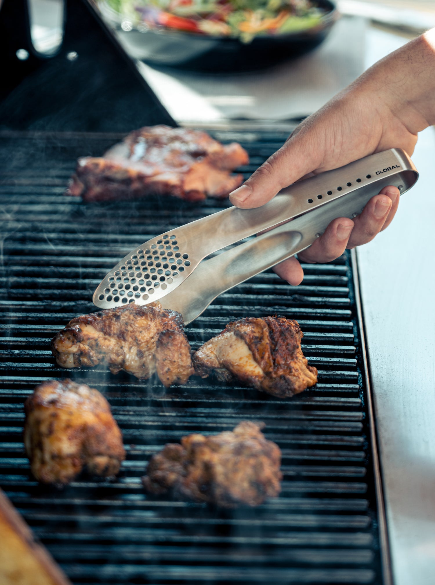 BBQ chef using Global stainless steel tongs to cook chicken on a grill with a salad in the background