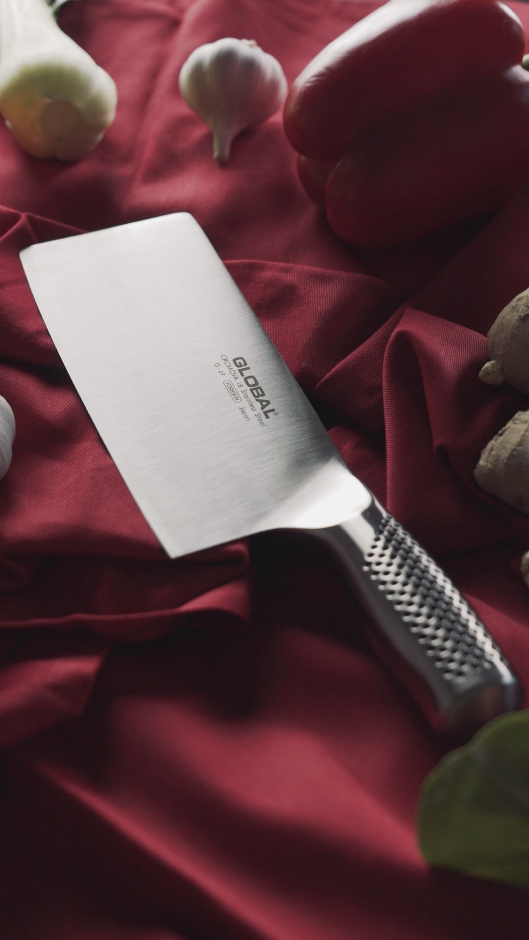 Close ups of Global Stainless steel Chinese' chef's knife with garlic, peppers and ginger on red silk.